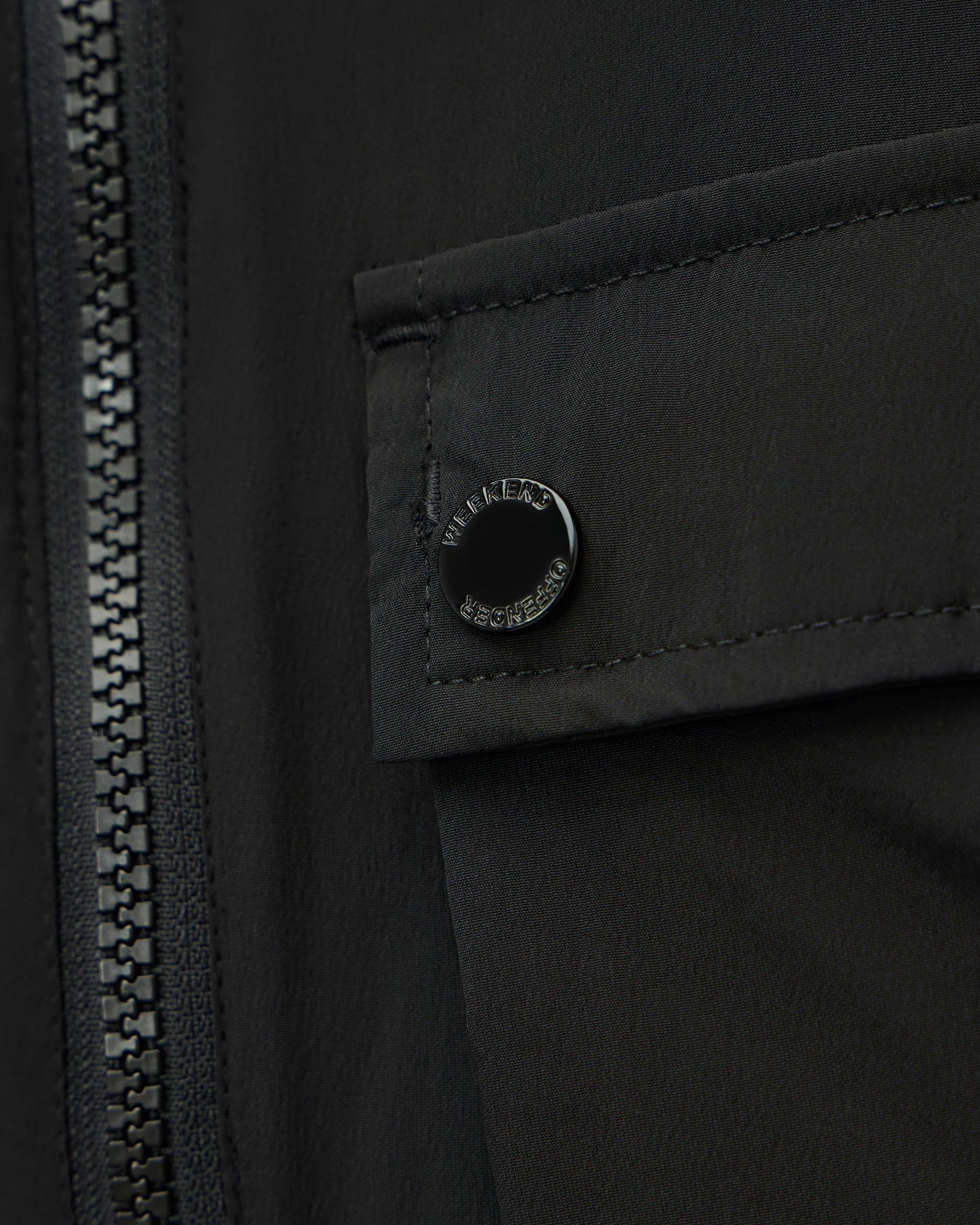 Close-up of a black jacket featuring a zippered front and a flap pocket with a circular black snap button engraved with 'MILITARY SPECIAL PURPOSE'.