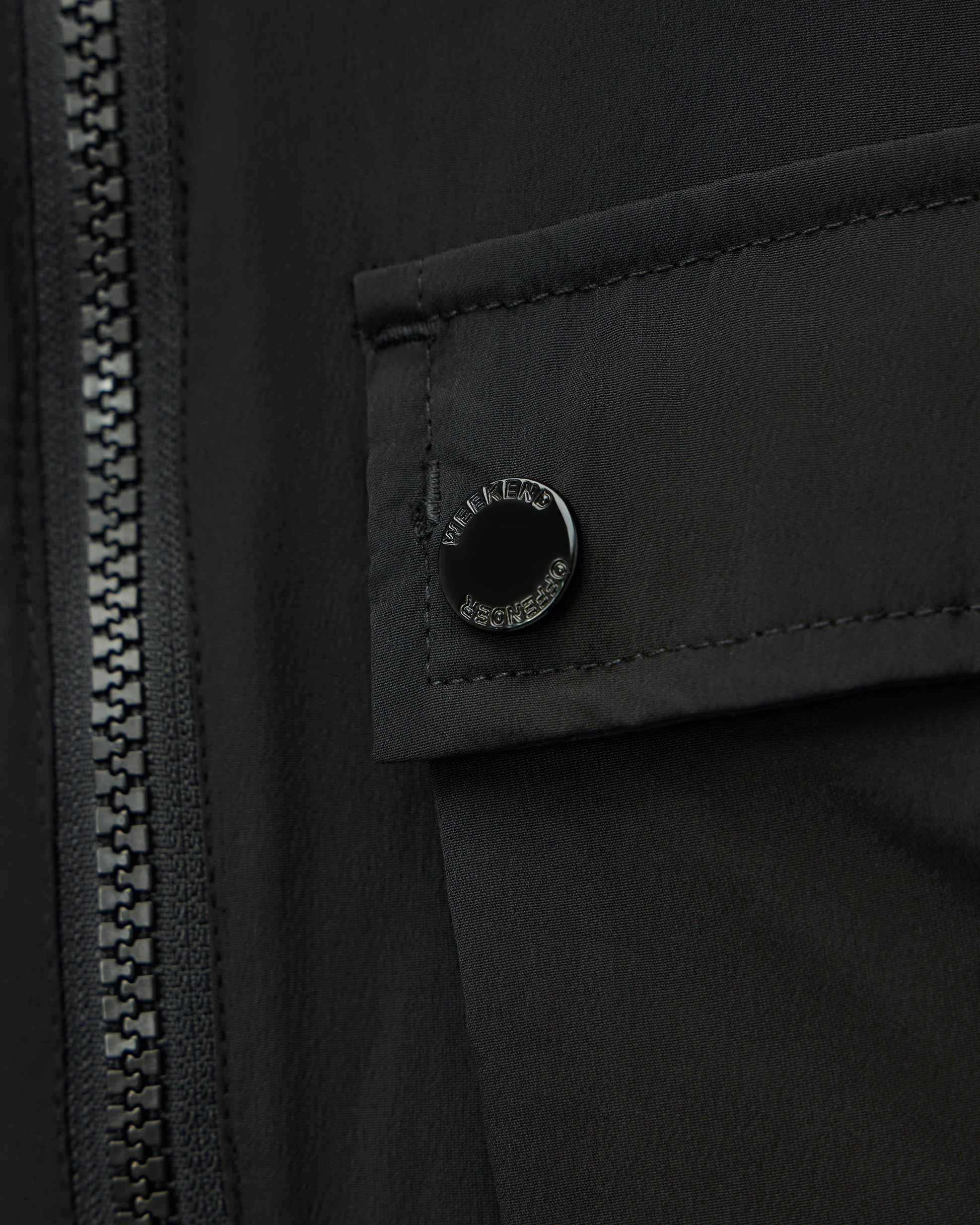 Close-up of a black jacket featuring a zippered front and a flap pocket with a circular black snap button engraved with 'MILITARY SPECIAL PURPOSE'.